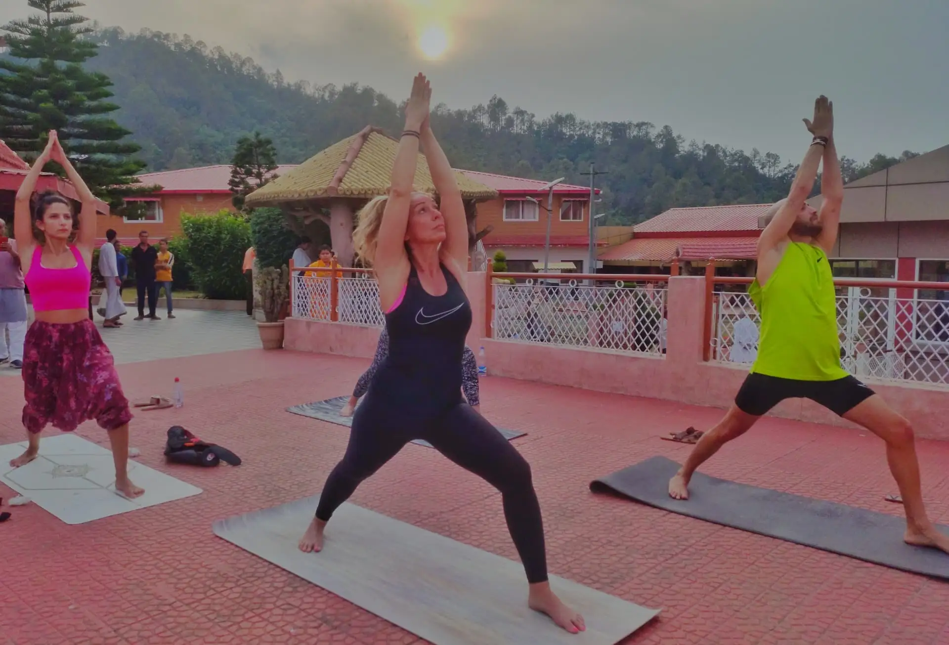 Practitioners in warrior pose on mats on the open ashram terrace at sunrise, with Kumaon hills behind